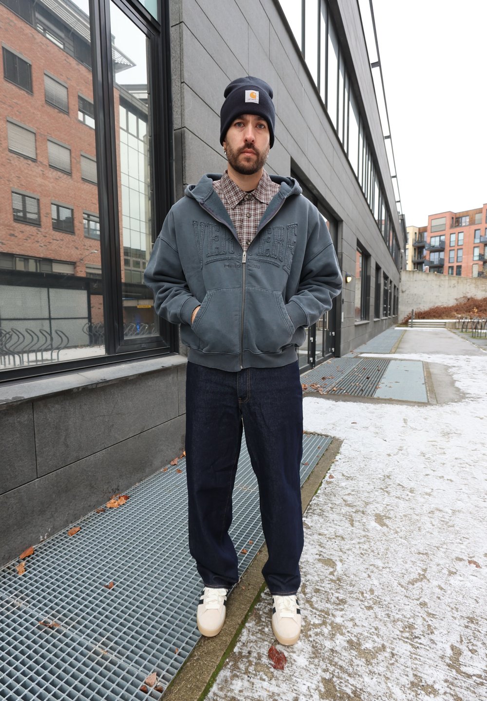 Mėlynas džemperis su užtrauktuku ir užrašu, po juo languota marškiniai, tamsios džinsinės kelnės ir šviesios spalvos sportbačiai. Lauko aplinka su sniegu ir pilkomis paviršių.