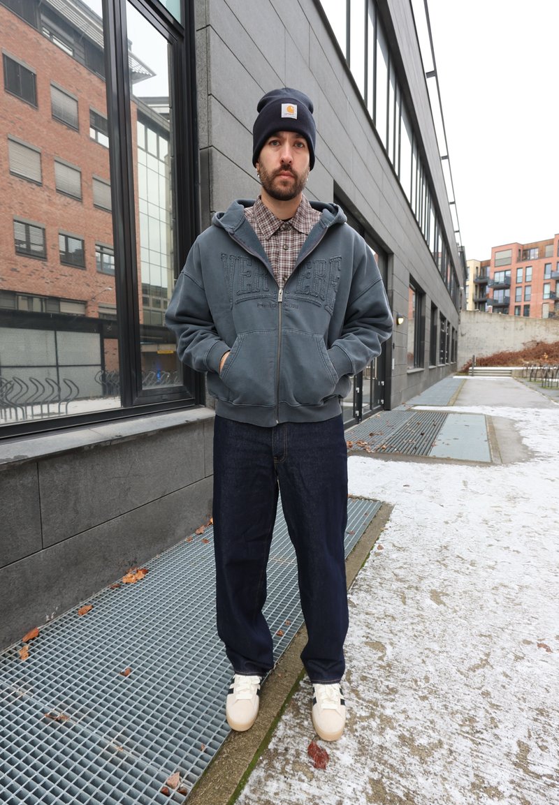Sweat à capuche zippé bleu avec texte, chemise à carreaux en dessous, pantalon en jean foncé et baskets de couleur claire. Environnement extérieur avec de la neige et des surfaces grises.