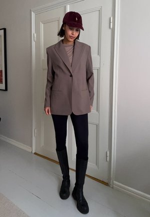 Mujer con una blazer marrón oversize, top malva, pantalones negros, botas negras y una gorra rojo oscuro, de pie en el interior contra una puerta blanca.