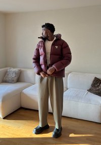 Homme en doudoune bordeaux en train de la fermer, portant un pantalon large beige et des chaussures noires, debout dans un salon lumineux avec des canapés blancs.