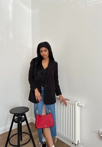 Sort blazer over en sort skjorte med knapper, parret med en denimnederdel med høj slids og en rød håndtaske. Har en baggrund med en træstol og en radiator.