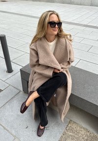 Beige fleece overcoat featuring a shawl collar, worn over a cream top. Black flared jeans paired with dark brown dress shoes. Seated outdoors on a stone bench.