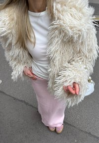 Casaco de lã com textura creme, blusa branca ajustada, saia de malha rosa e sapatos rosa claro. Pequena mala branca de forma arredondada com detalhes em dourado.