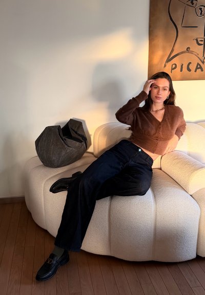 Mujer con cárdigan marrón y pantalones negros sentada en un sofá blanco curvo, tocándose el cabello, con un bolso oscuro a su lado y arte en la pared.