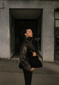 Giacca di pelle nera con colletto alto, abbinata a una borsa in velluto nera. Modella in piedi contro uno sfondo di un edificio moderno in pietra.