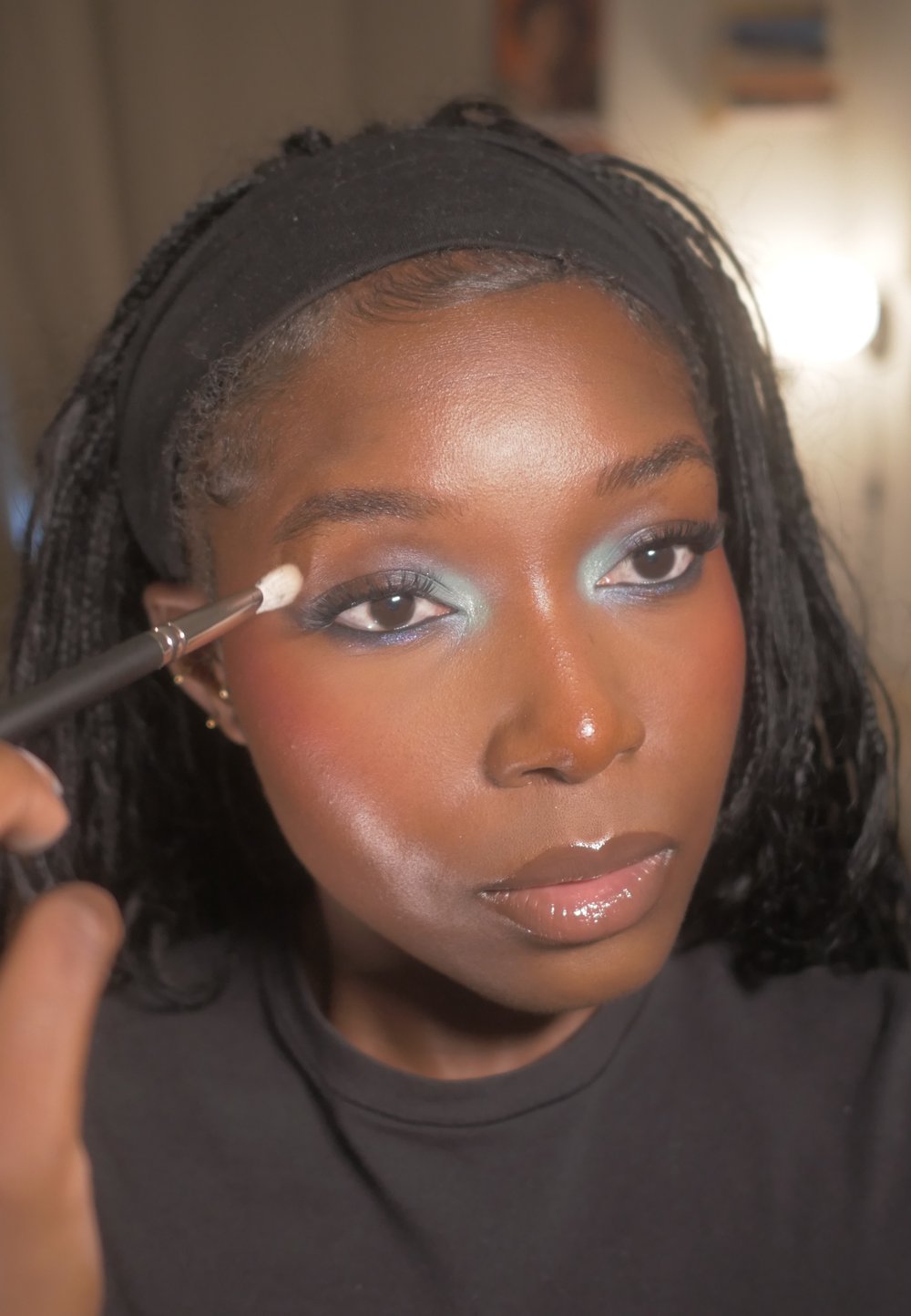 Application de maquillage avec un bandeau noir, mettant en avant un fard à paupières bleu, du blush et des lèvres brillantes. Vue en gros plan, focus sur les traits du visage et les outils de maquillage.