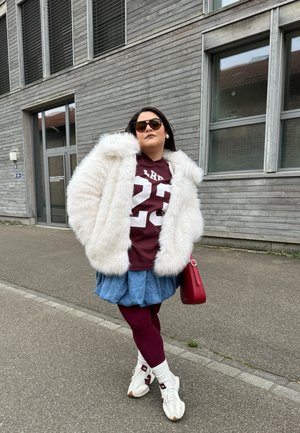 Abrigo de pelaje blanco y esponjoso, camisa burdeos con el número 23, falda de mezclilla, mallas burdeos y zapatillas blancas con acentos rojos. Bolsito rojo.