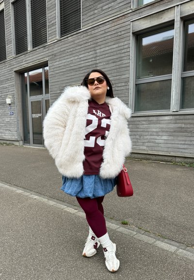 Abrigo de pelaje blanco y esponjoso, camisa burdeos con el número 23, falda de mezclilla, mallas burdeos y zapatillas blancas con acentos rojos. Bolsito rojo.