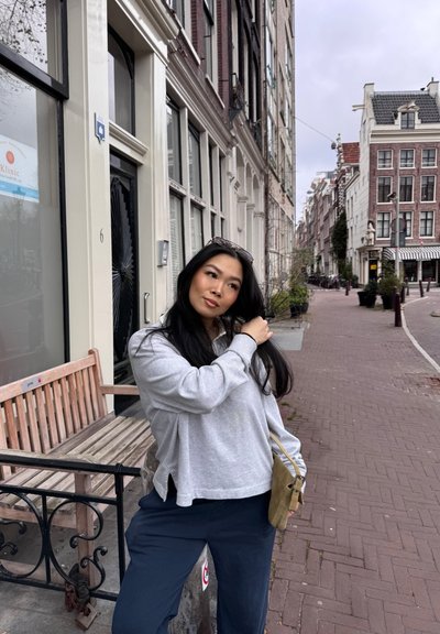 Sudadera gris claro y pantalones oscuros azules. Cabello largo y negro con gafas de sol en la cabeza. Calle urbana con un banco de madera y pavimento de ladrillo.