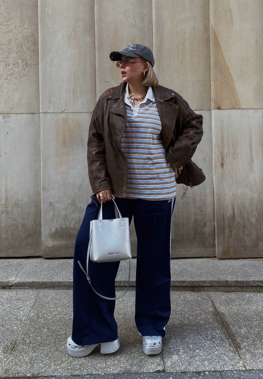 Καφέ δερμάτινο μπουφάν πάνω από ένα ριγέ πόλο, ναυτικές φόρμες, ασημένια τσάντα και παπούτσια με πλατφόρμα. Ένα γκρίζο καπέλο και γυαλιά ολοκληρώνουν την εμφάνιση.