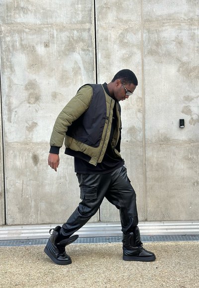 Chaqueta tipo puffer con secciones en verde oliva y negro, pantalones cargo negros y zapatillas altas negras, con un fondo de hormigón.