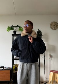 Veste bomber noire avec col en fausse fourrure, fermeture éclair et deux poches avant ; associée à un pantalon de jogging plissé gris clair.