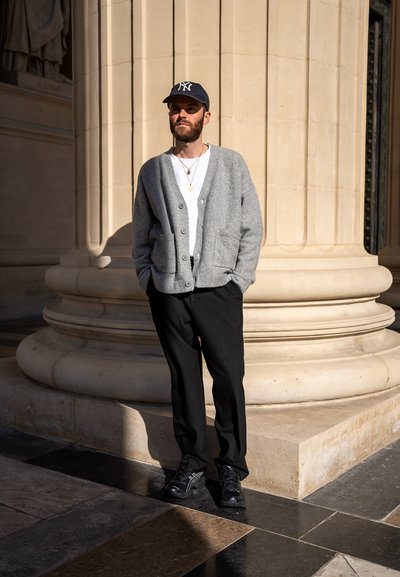 Cárdigan gris con botones y bolsillos frontales, camiseta blanca, pantalones negros y zapatillas negras. De pie cerca de una columna de piedra.