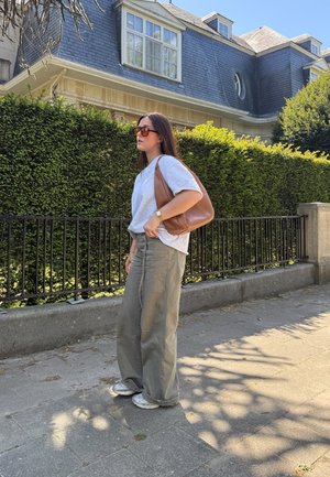 Camiseta gris claro, pantalones anchos oliva y zapatillas plateadas. Bolso marrón de hombro, gafas de sol rectangulares, de pie contra un seto verde.