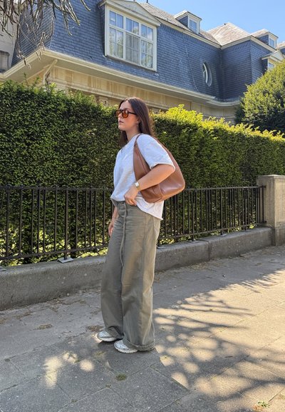 Camiseta gris claro, pantalones anchos oliva y zapatillas plateadas. Bolso marrón de hombro, gafas de sol rectangulares, de pie contra un seto verde.