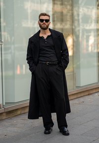 Černý dlouhý kabát přes černou košili a kalhoty. Oblečení obsahuje elegantní boty a sluneční brýle, které jsou umístěny na pozadí moderní skleněné architektury.