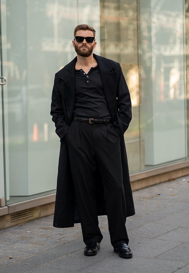 Casaco longo preto sobre uma camisa e calças pretas. O conjunto inclui sapatos elegantes e óculos de sol, tendo como fundo uma moderna parede de vidro.