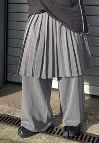 Sivá plisovaná sukňa cez široké sivé nohavice; látka pôsobí textúrovaným dojmom. Čierne topánky s hrubou podrážkou. Jednoduchý čierny top s kontrastným prešitím.