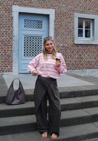 Helles rosa gestreiftes Sweatshirt, weite schwarze Hose mit Drapierung, braune Handtasche und Flip-Flops, mit einem To-Go-Kaffeebecher in der Hand.