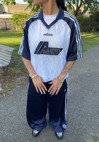 Adidas sporta tērps, kas sastāv no balta krekla ar melnu logo, tumši ziliem biksēm ar baltām svītrām un melniem čību apaviem ar grafisku dizainu.