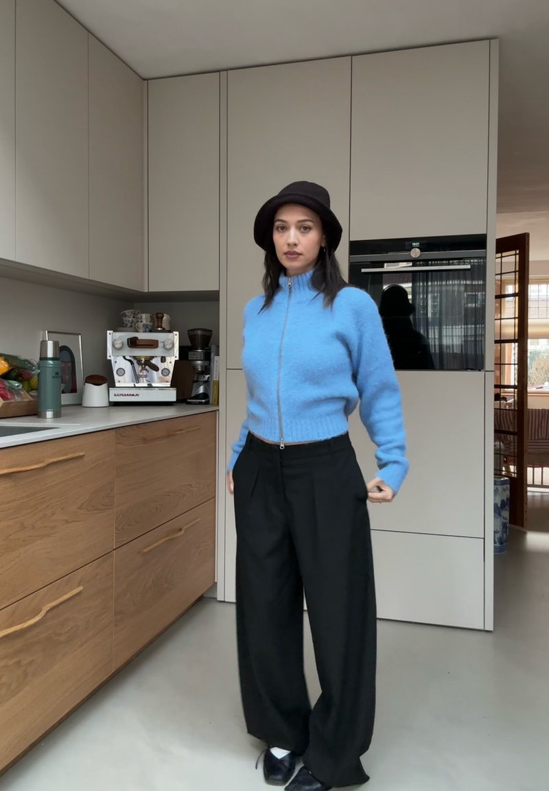 Pull à fermeture éclair bleu clair en tissu duveteux, pantalon noir à jambes larges, chapeau noir ; fond de cuisine avec armoires en bois et appareils électroménagers.