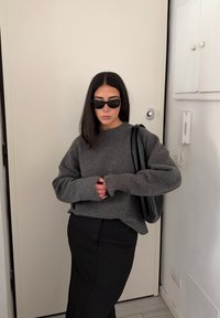 Pull gris en maille à manches longues et col rond, associé à une jupe crayon noire. Les accessoires comprennent des lunettes de soleil foncées et un sac noir.
