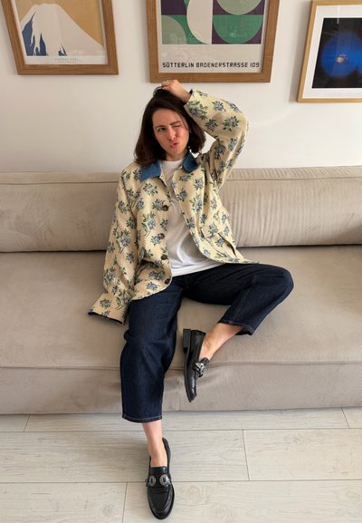 Mujer sentada en un sofá beige, vistiendo una chaqueta floral, camisa blanca, jeans oscuros y mocasines negros, con una obra de arte enmarcada en la pared detrás de ella.