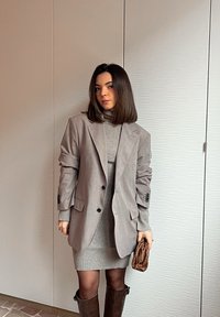 Femme debout à l'intérieur, portant un blazer gris et une robe en maille assortie, tenant une petite pochette tissée marron, avec des bottes marron montant jusqu'aux genoux.