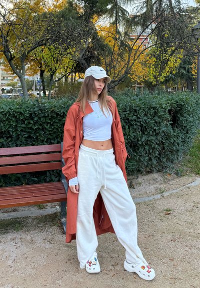 Un largo abrigo naranja sobre un top blanco corto, pantalones blancos holgados y zuecos blancos con decoraciones coloridas. El fondo presenta arbustos verdes.