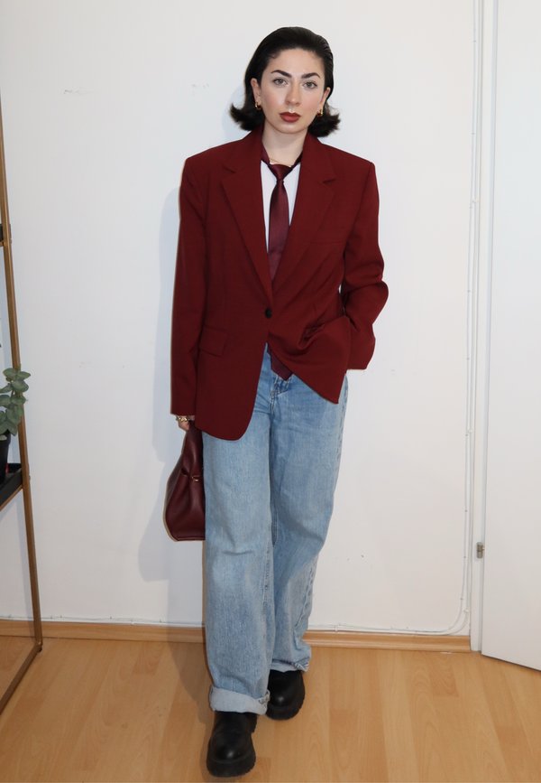 Woman wearing a burgundy blazer, matching tie, light blue jeans, black boots, and holding a burgundy handbag standing indoors on a wooden floor.
