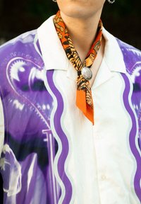Short-sleeved shirt in white with purple graphic designs, paired with an orange and black patterned neck scarf featuring a silver clasp.
