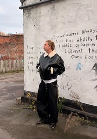 Personne aux cheveux gominés portant une veste en cuir noire avec un col en peau lainée et un pantalon large noir, debout contre un mur couvert de graffitis.