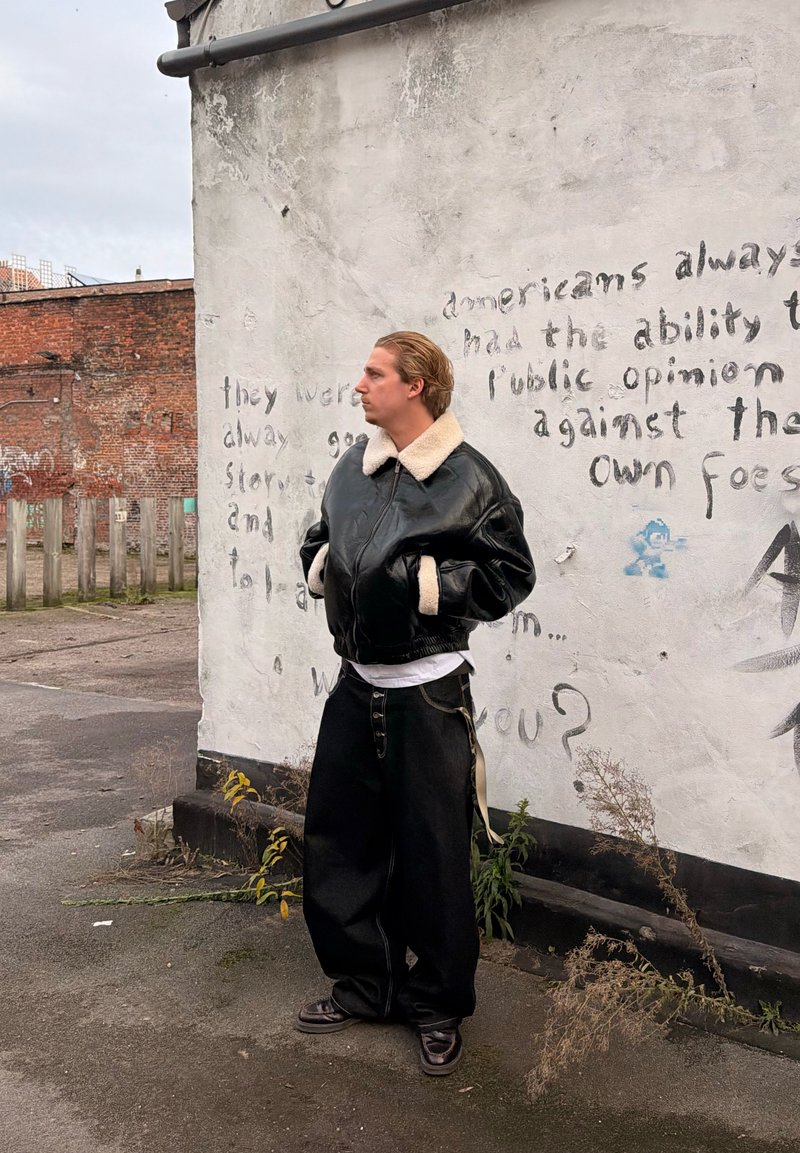 Personne aux cheveux gominés portant une veste en cuir noire avec un col en peau lainée et un pantalon large noir, debout contre un mur couvert de graffitis.