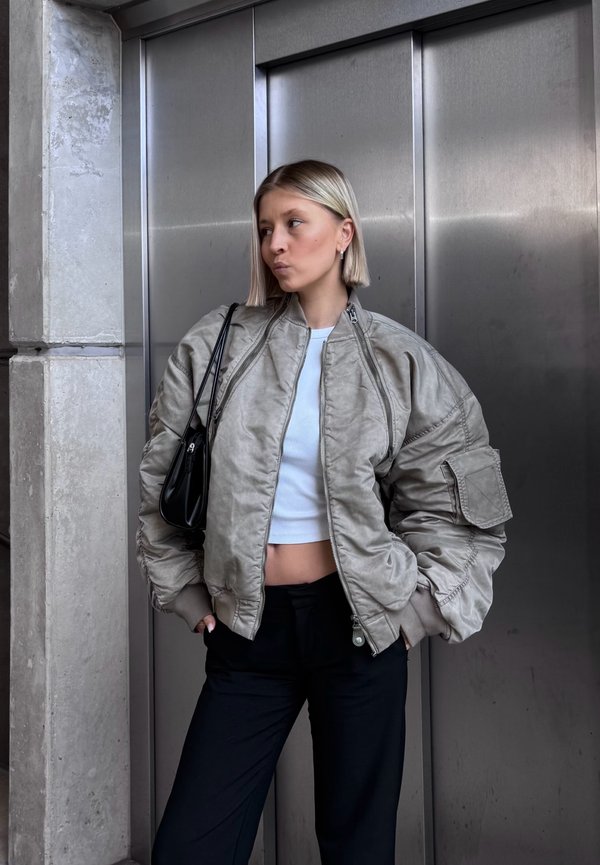 Vadderad bomberjacka i ljusgrått, med hög krage, ribbade ärmslut och sidofickor, tillsammans med en kort vit topp och svarta byxor.