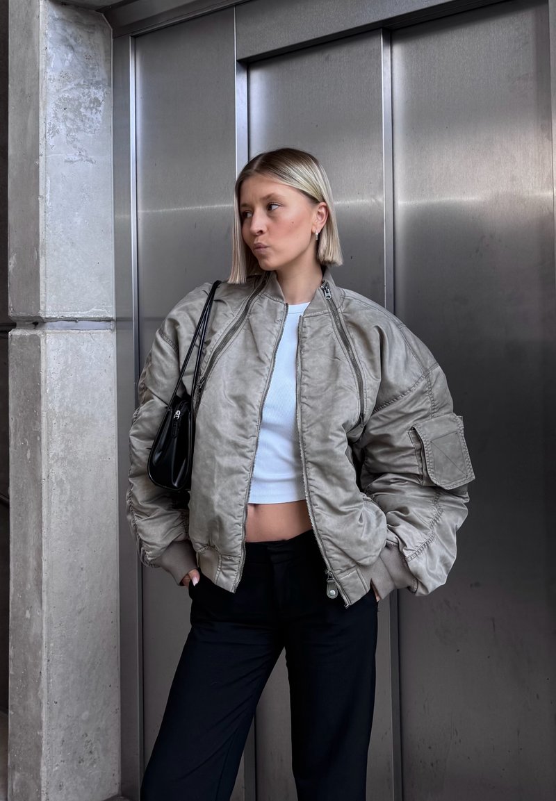 Dicke Bomberjacke in Hellgrau mit hohem Kragen, gerippten Bündchen und seitlichen Taschen, kombiniert mit einem kurzes weißen Top und schwarzen Hosen.