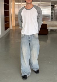 Mladá osoba v bílo-šedém tričku s dlouhým rukávem, volných světle modrých džínách a černých botách stojí uvnitř s rukama za zády.