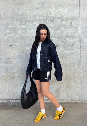 Mujer joven con chaqueta vaquera, pantalones cortos negros Adidas y zapatillas amarillas, sosteniendo un bolso grande negro con textura, posando frente a una pared de hormigón.