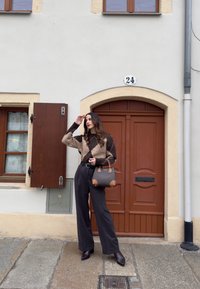 Femme en pull marron à losanges et pantalon noir tenant un sac à main, debout sur un pavé en pierre devant un bâtiment beige avec une porte marron numérotée 24.