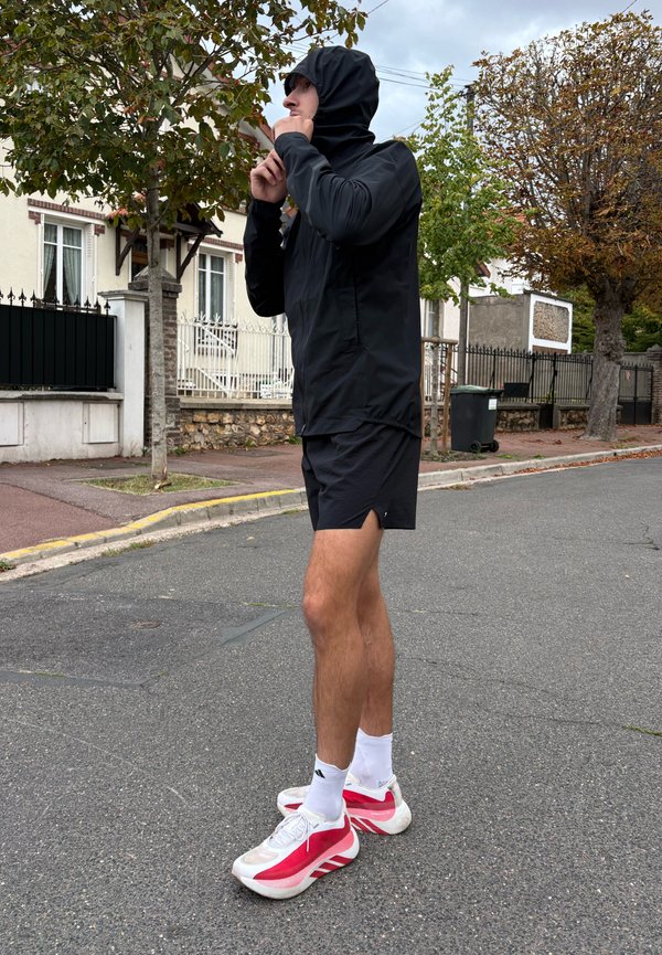 Une personne portant un haut noir à capuche et un short ajuste sa capuche dans une rue résidentielle. Elle porte des chaussettes blanches et des chaussures de course roses.