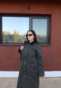 Donna con un lungo cappotto scuro e occhiali da sole neri in piedi contro un muro di mattoni rossi con una finestra che riflette alberi e cielo.