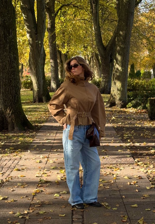 Une femme portant des lunettes de soleil, une veste marron ceinturée et un jean ample pose sur un sentier jonché de feuilles, sous des arbres d’automne.