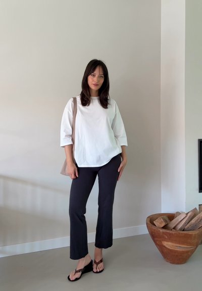 Camisa de algodón blanca de gran tamaño con mangas de 3/4, combinada con pantalones oscuros de campana y chanclas marrones, de pie contra una pared gris claro.