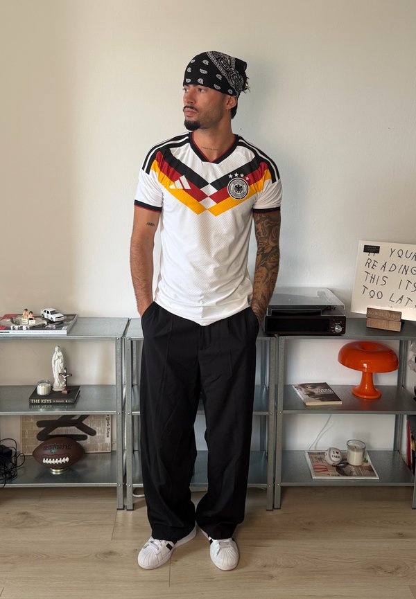 Man met tatoeages die een Duits voetbalshirt en een bandana draagt, staat met zijn handen in zijn zakken in een kamer met planken vol boeken en decoratie.