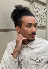Veste en denim beige clair avec des fermetures à boutons et des détails lacés sur les côtés. Le sujet a des cheveux bouclés et porte une boucle d'oreille en argent.