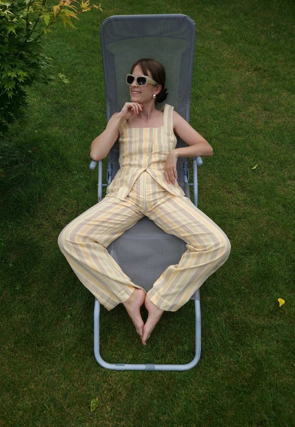 Grijze ligstoel met een mesh-stof, omringd door groen gras. Geel en wit gestreept outfit, met een knoopjesshirt en brede broek.