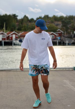 Camisa blanca de manga corta con texto azul, pantalones cortos de baño con un colorido patrón de palmeras y zapatos azul claro. El fondo presenta estructuras de madera junto al agua.