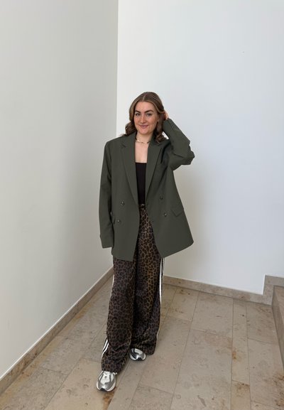 Mujer de pie en el interior con un blazer oversized color oliva, top negro, pantalones con estampado de leopardo y zapatillas plateadas, sonriendo con una mano tocándose el pelo.
