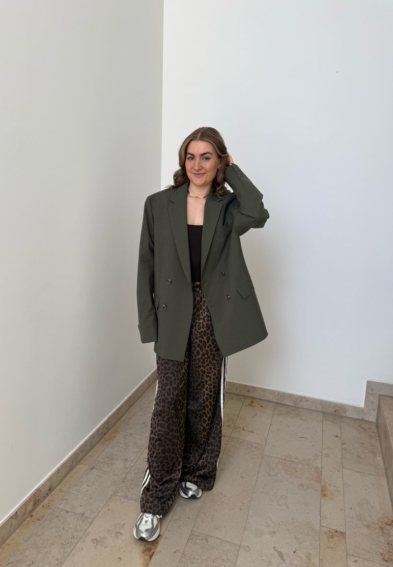 Donna in piedi all'interno, indossa un blazer oversize oliva, top nero, pantaloni con stampa leopardata e sneakers argento, sorride con una mano che tocca i capelli.