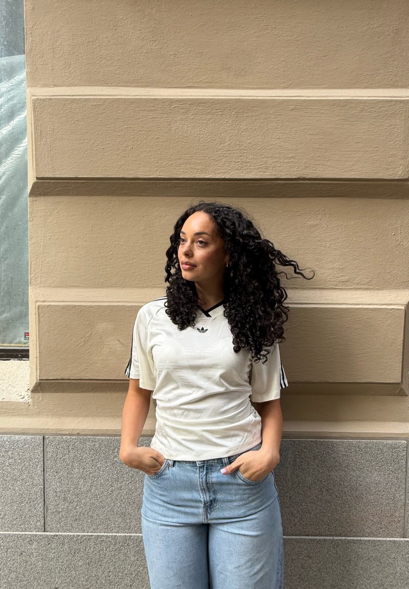 Femme aux cheveux bouclés portant une chemise blanche et un jean bleu, debout contre un mur beige, les mains dans les poches, regardant sur le côté.