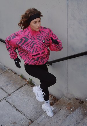 Cortavientos estampado en rosa y gris con texto "NIKE AIR", leggings negros, zapatillas blancas y guantes, en una pose activa de carrera en unas escaleras.